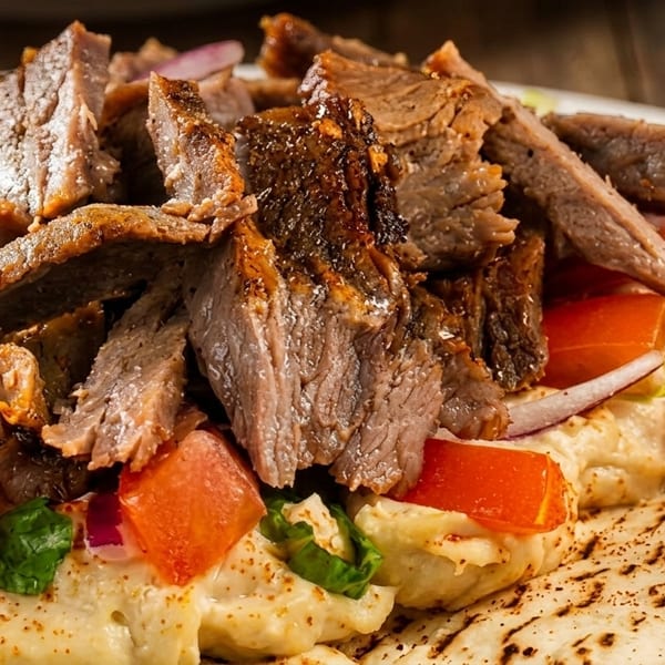 Close-up of a beef shawarma plate with hummus, tomatoes, onions, and pickled turnips on pita bread.